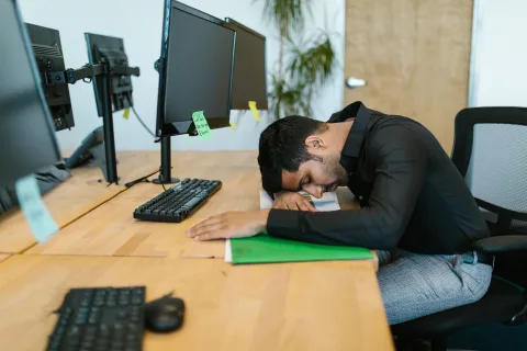 Un homme assis à son bureau de travail, la tête posée sur ses bras croisés pour une courte sieste.