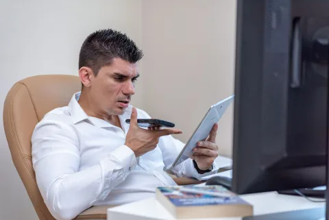 Un jeune homme en chemise parle devant son smartphone posé sur un bureau en bois