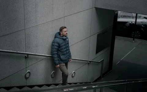 Un homme marchant dans une rue pavée en ville sous une fine pluie en portant une veste technique verte sur un pantalon chino sombre.