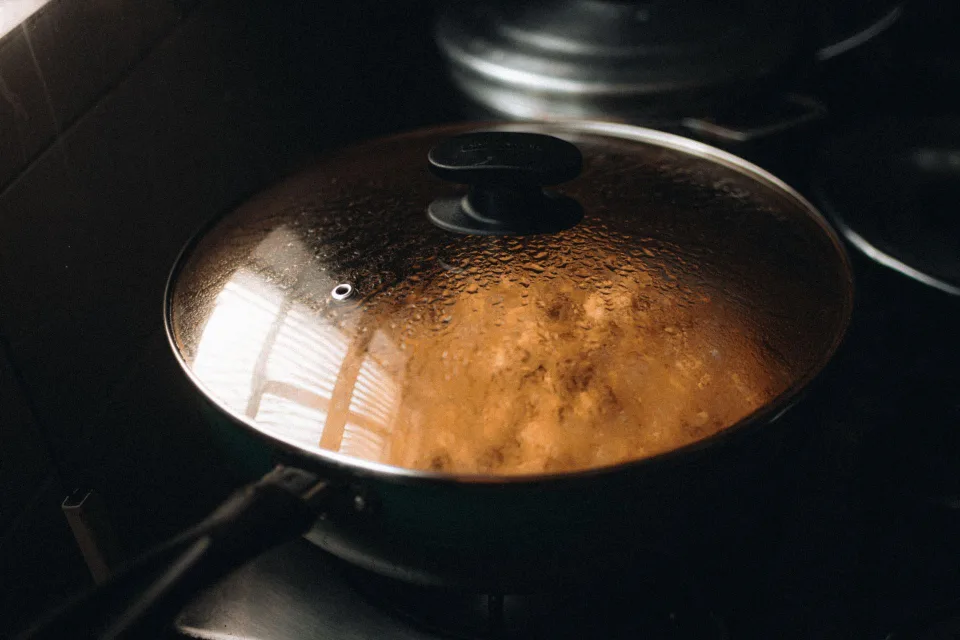 Une casserole en inox fermée par un couvercle transparent laissant échapper de la vapeur sur une plaque de cuisson éteinte.