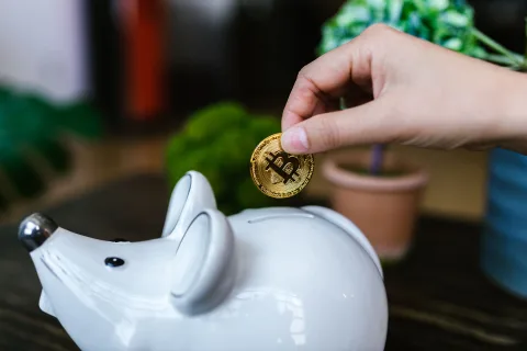 Une main glissant des pièces de monnaie dans une tirelire rose en forme de cochon posée sur une table en bois clair.