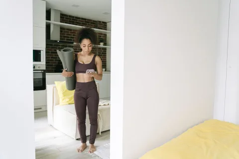 Une jeune femme en tenue de sport réalise des fentes dans son salon en suivant les instructions sur son smartphone posé au sol.