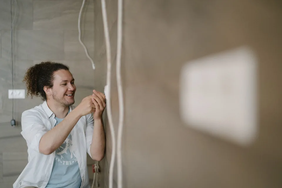 Main d'un électricien insérant un petit boîtier bleu derrière un interrupteur mural démonté.