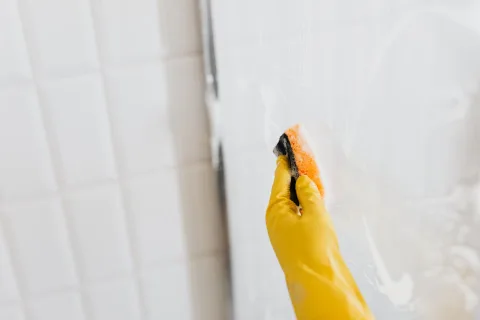 Une main vaporisant un produit naturel sur une paroi de douche transparente couverte de calcaire.