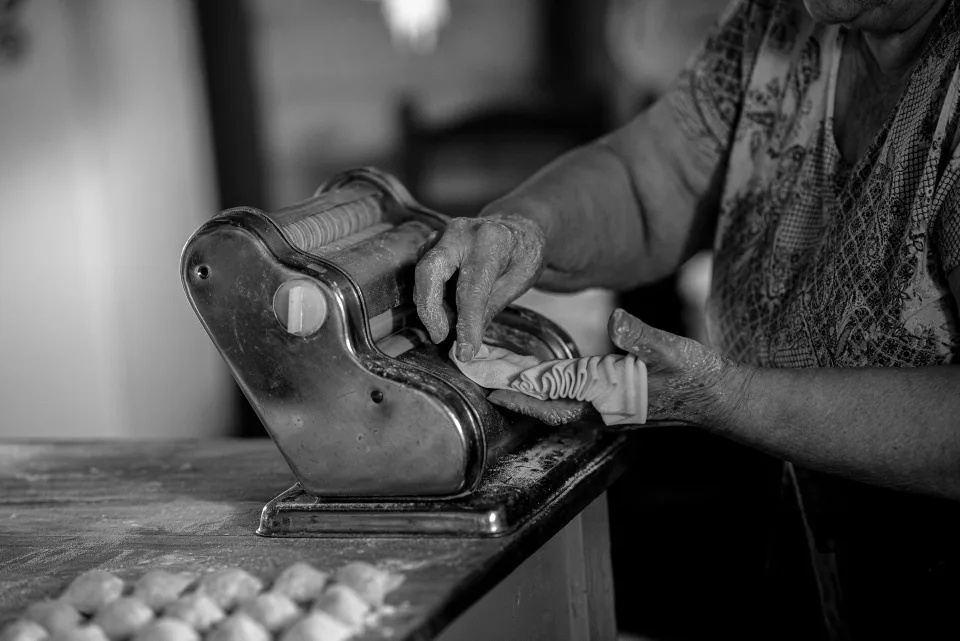 Une femme âgée souriante préparant des pâtes fraîches dans une cuisine ensoleillée.