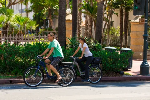 Un jeune couple roule à vélo sur une piste cyclable bordée d'arbres le long d'un lac tranquille.