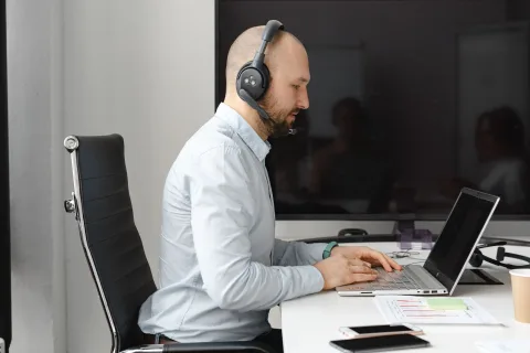 Une personne de dos portant un casque audio sans fil travaille sur un ordinateur portable dans un bureau lumineux.