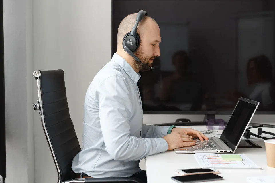 Une personne de dos portant un casque audio sans fil travaille sur un ordinateur portable dans un bureau lumineux.