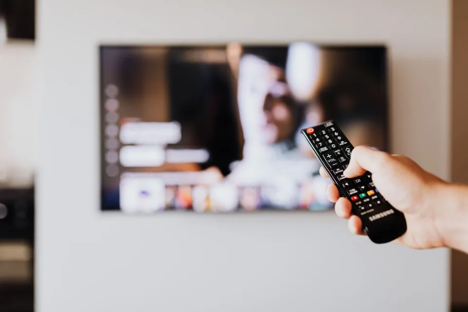 Une personne assise sur son canapé tient une télécommande devant un écran de télévision affichant un menu de streaming.