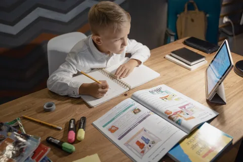 Un enfant étudie devant un ordinateur avec un cahier et un stylo à la main.